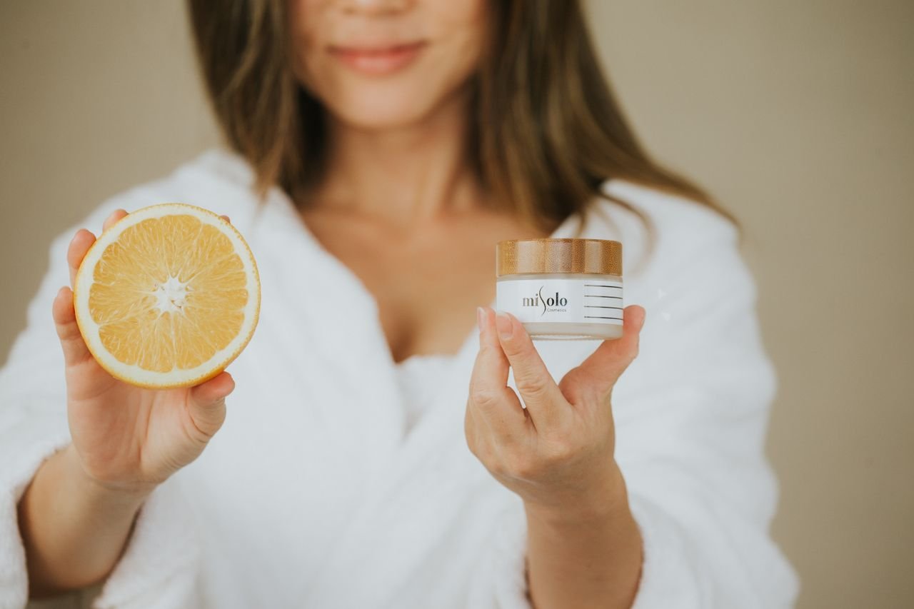 About A woman in a robe holding skincare cream and an orange slice, highlighting natural beauty and freshness.