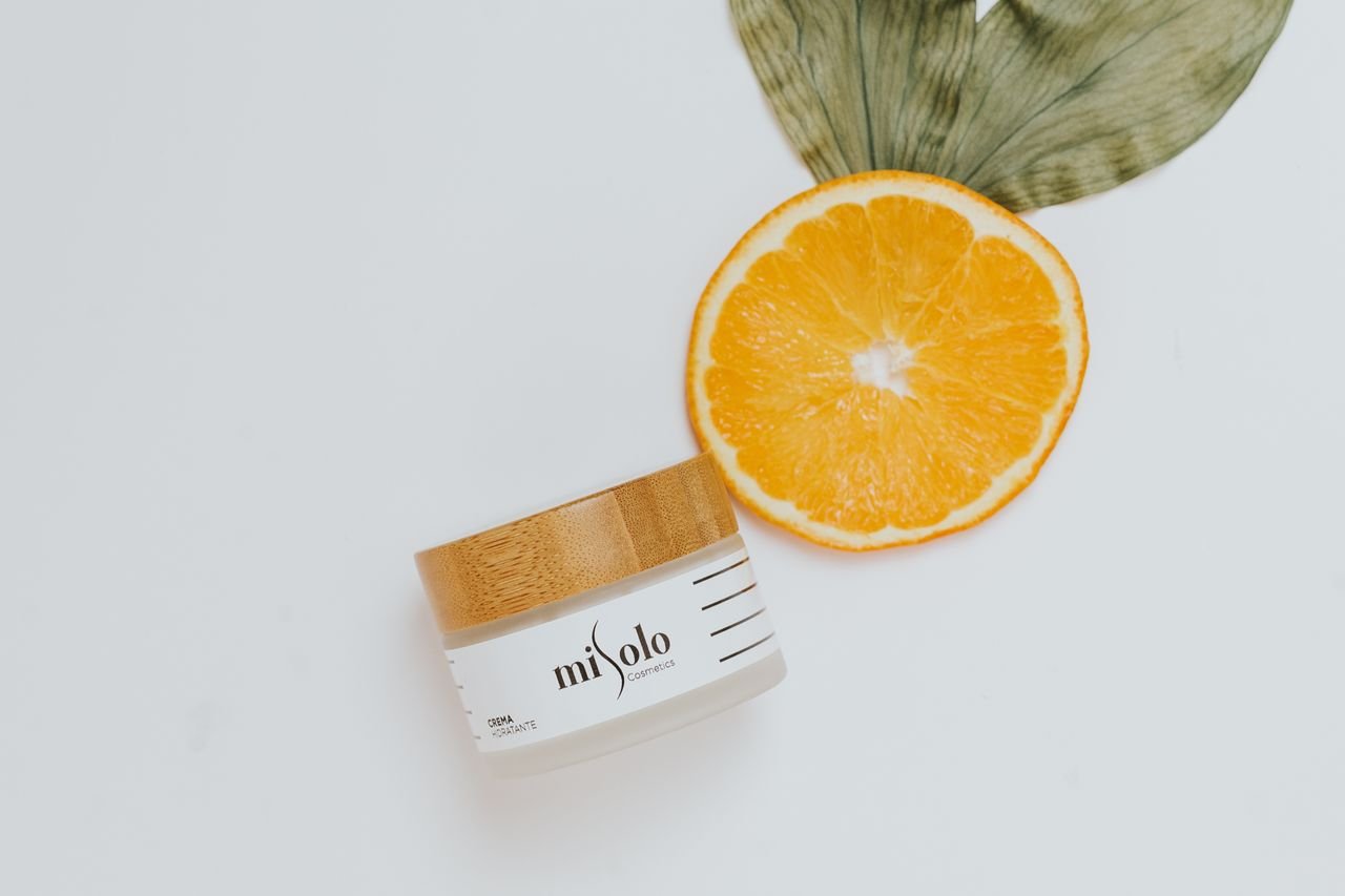 About Close-up of orange slice and skincare jar with leaf on white background.